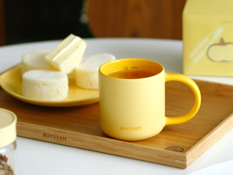 Żółty kubek ceramiczny z napisem \'BUYDEEM\' stoi na drewnianej tacy obok talerza z białymi ciastkami. W tle widoczny jest żółty karton z grafiką owocu.