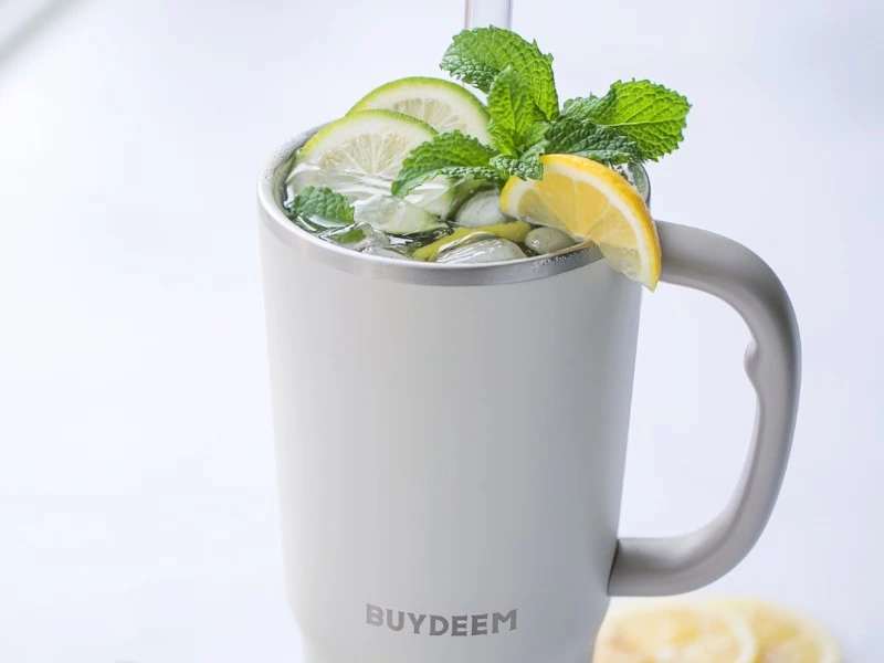 Kubek termiczny w kolorze białym z logo \'BUYDEEM\', wypełniony napojem z plasterkami cytryny, limonki i liśćmi mięty, ze słomką.
