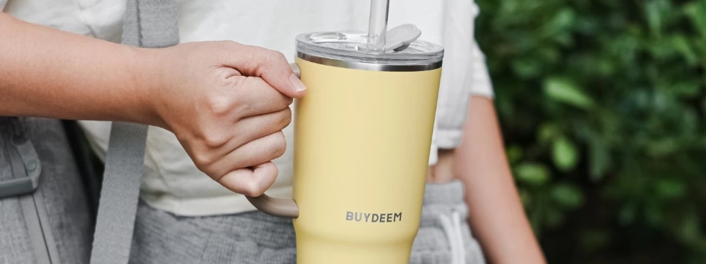 Osoba trzyma żółty kubek termiczny z przezroczystą słomką, na którym widnieje napis \'BUYDEEM\'. W tle widoczne są zielone liście.