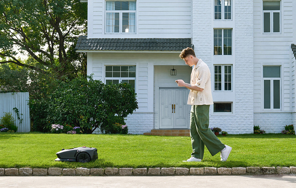Robot koszący DREAME Mova 600 robot na trawie za nim osoba z telefonem w dłoni w tle dom i rośliny wyznacza wirtualne granice aplikacja MOVAHome wielostrefowe mapy harmonogramy pracy wyłączać obszary kontrolę koszenia bezpośrednio z poziomu telefonu bezpieczeństwo Twoich kwiatów wbudowany moduł Wi-Fi