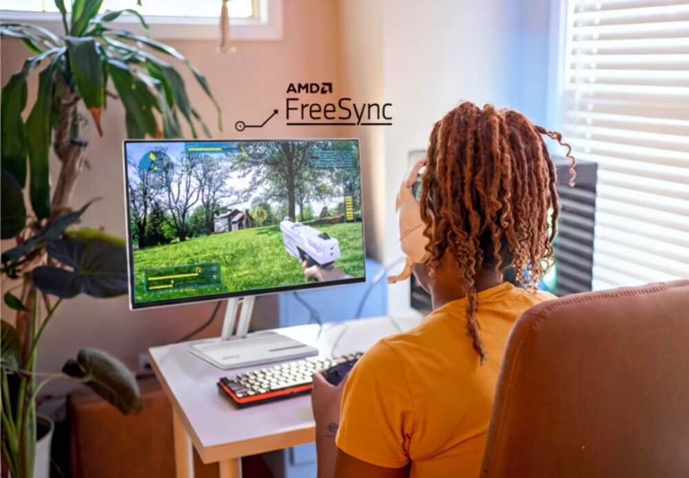 Osoba siedzi przy biurku, grając w grę na monitorze, na którym widoczny jest krajobraz z drzewami i budynkiem. Na ekranie znajduje się napis \'AMD FreeSync\'.