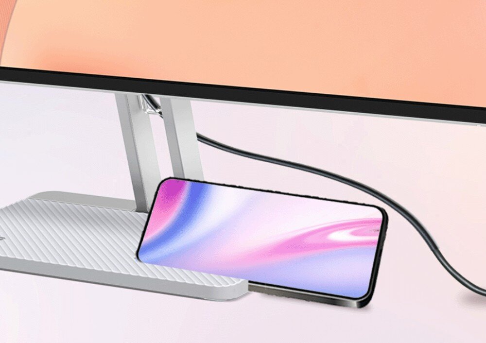 Na obrazie widoczny jest fragment monitora z metalową podstawą oraz smartfon z gradientowym ekranem opartym o podstawę. Kabel jest podłączony do monitora.