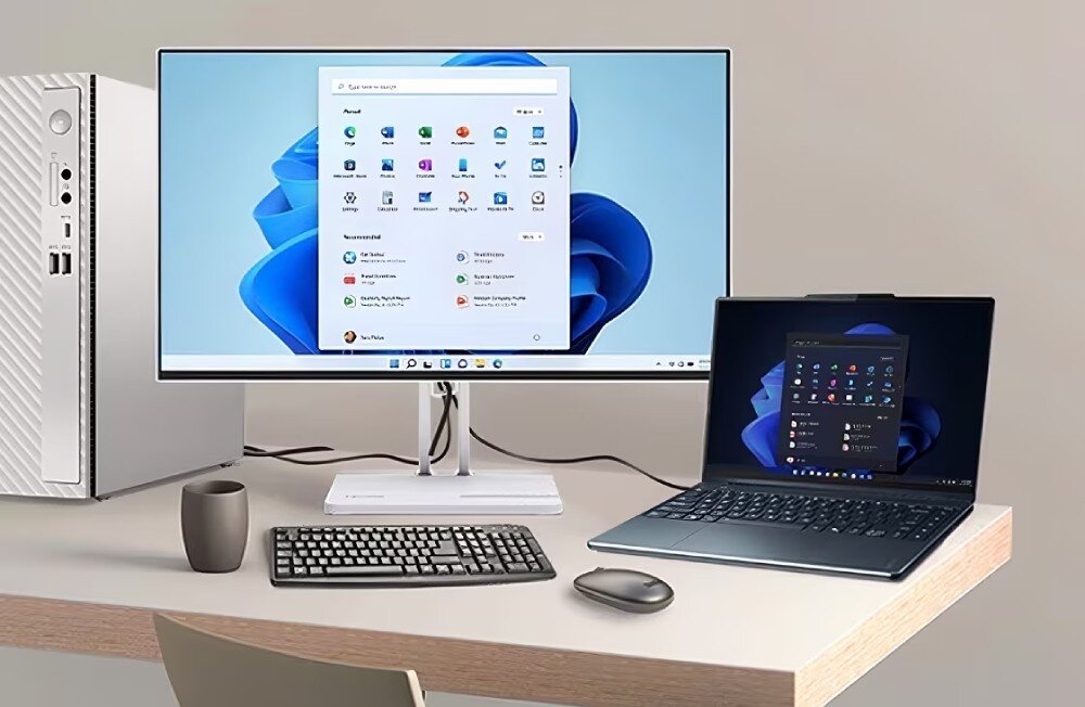 Na biurku znajduje się monitor z wyświetlonym ekranem startowym systemu operacyjnego, obok laptop z podobnym ekranem. Obok monitora stoi komputer stacjonarny, klawiatura, myszka i kubek.
