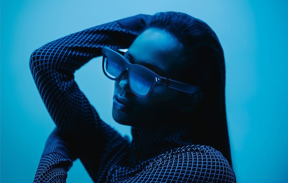 Osoba nosi okulary w futurystycznym stylu, w otoczeniu niebieskiego oświetlenia. Ubrana jest w wzorzystą odzież z długim rękawem.