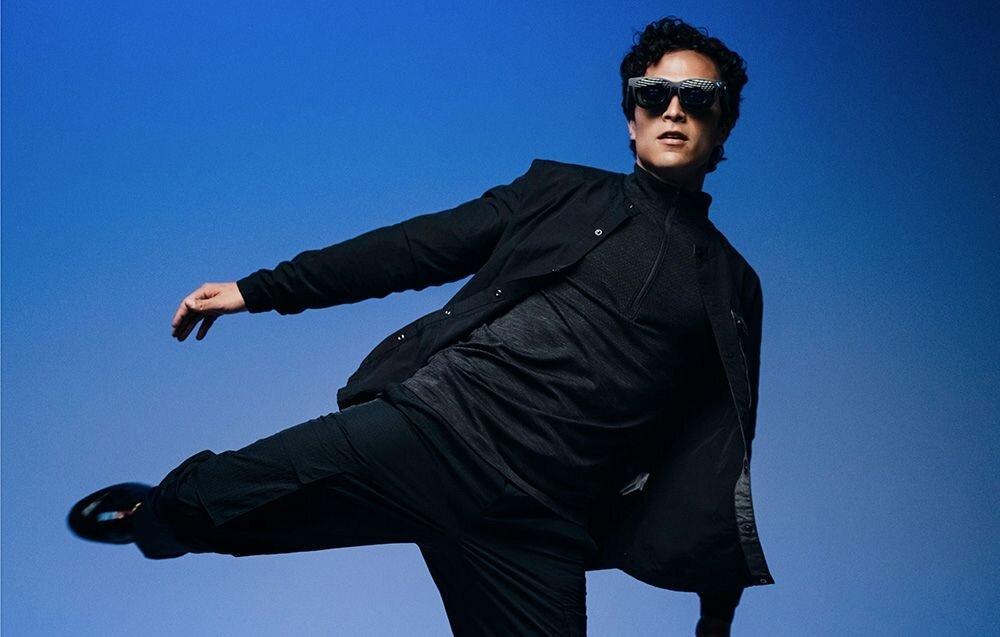 Osoba w ciemnym ubraniu i okularach przeciwsłonecznych wykonuje dynamiczny ruch na tle niebieskiego gradientu.