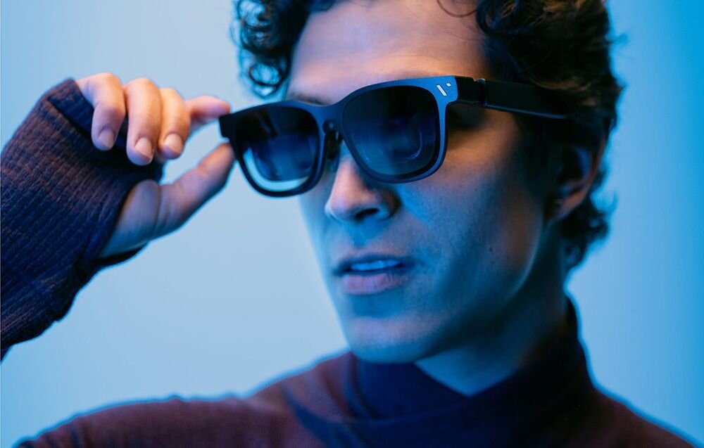 Osoba nosi ciemne okulary przeciwsłoneczne w nowoczesnym stylu. Tło jest w odcieniach niebieskiego, co nadaje zdjęciu chłodny ton.