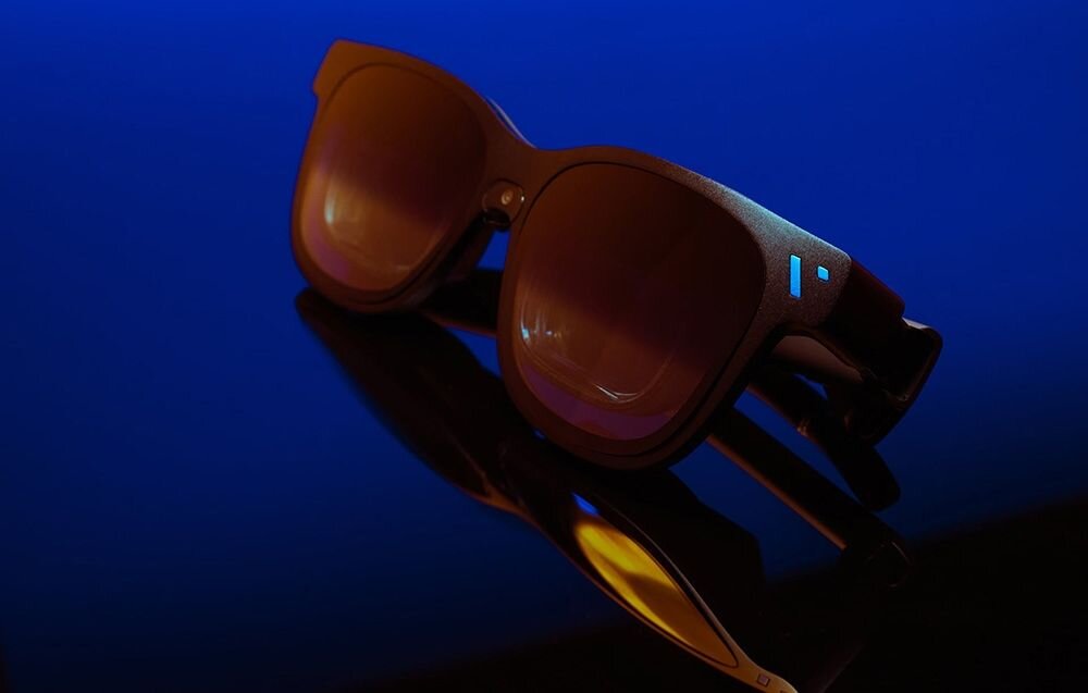 Okulary przeciwsłoneczne z ciemnymi szkłami i oprawkami, leżące na powierzchni odbijającej światło. Tło jest w odcieniach niebieskiego, co podkreśla nowoczesny design produktu.