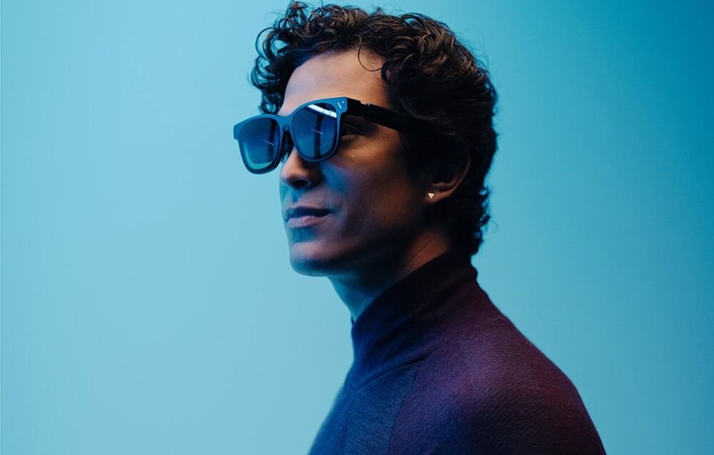 Osoba nosi nowoczesne okulary z ciemnymi szkłami na tle niebieskiego światła. Okulary mają masywną oprawkę.