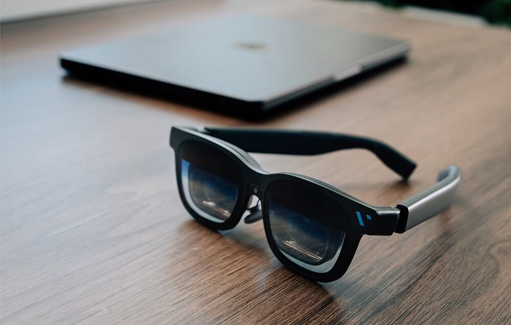 Na drewnianym stole leżą czarne okulary z grubymi oprawkami, a w tle znajduje się zamknięty laptop. Okulary mają niebieski akcent na zausznikach.