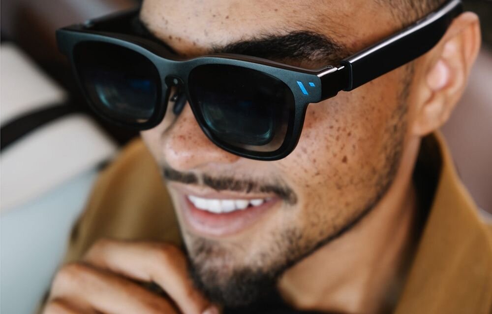 Osoba nosi czarne okulary z grubymi oprawkami, na których widoczne jest niebieskie logo. Okulary mają nowoczesny design i ciemne soczewki.