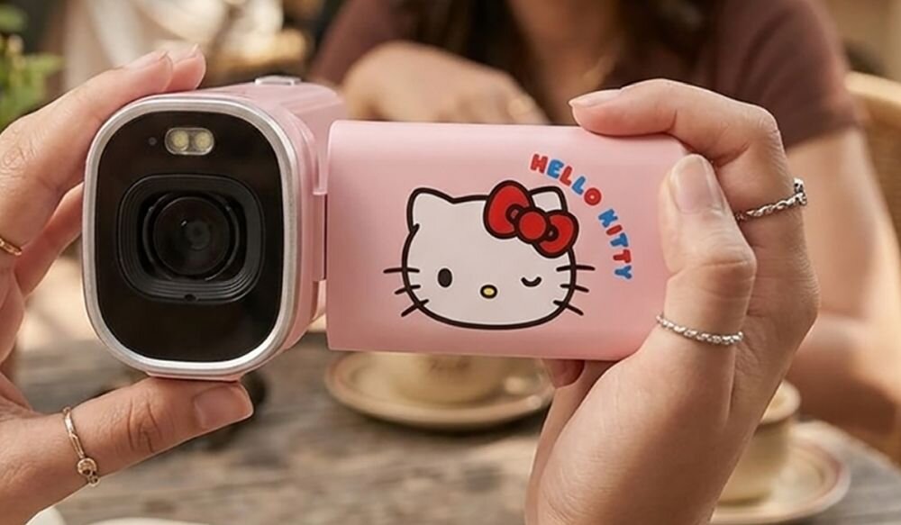 Różowa kamera cyfrowa z motywem Hello Kitty, trzymana w dłoniach. Na obudowie widoczny jest napis \'HELLO KITTY\' oraz wizerunek postaci Hello Kitty.
