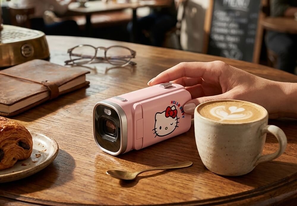 Różowa kamera cyfrowa z motywem Hello Kitty leży na drewnianym stole obok filiżanki kawy i rogalika. Obok kamery znajduje się para okularów i notes.
