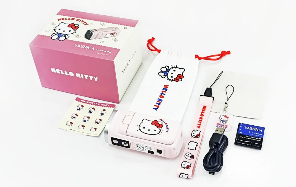 Zestaw zawiera różową kamerę cyfrową z motywem Hello Kitty, pudełko, etui, smycz, kabel USB, baterię oraz naklejki. Na pudełku i etui widnieje napis \'HELLO KITTY\'.
