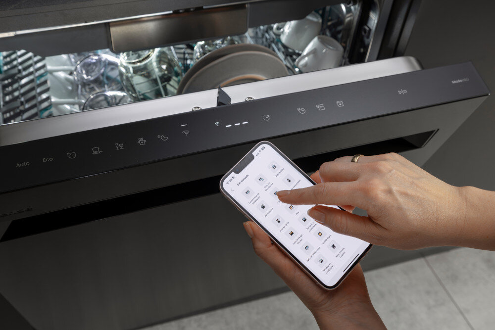 ZMYWARKA HISENSE HS693A90XAD  Na zdjęciu widać otwartą zmywarkę z panelem sterowania w czarnym wykończeniu, a przed nią osoba trzyma smartfon z uruchomioną aplikacją do obsługi urządzenia. Jedna dłoń wybiera ustawienia programu na ekranie telefonu, co podkreśla możliwość zdalnego sterowania zmywarką wifi zmywarka connectlife sterowanie smartfonem aplikacja