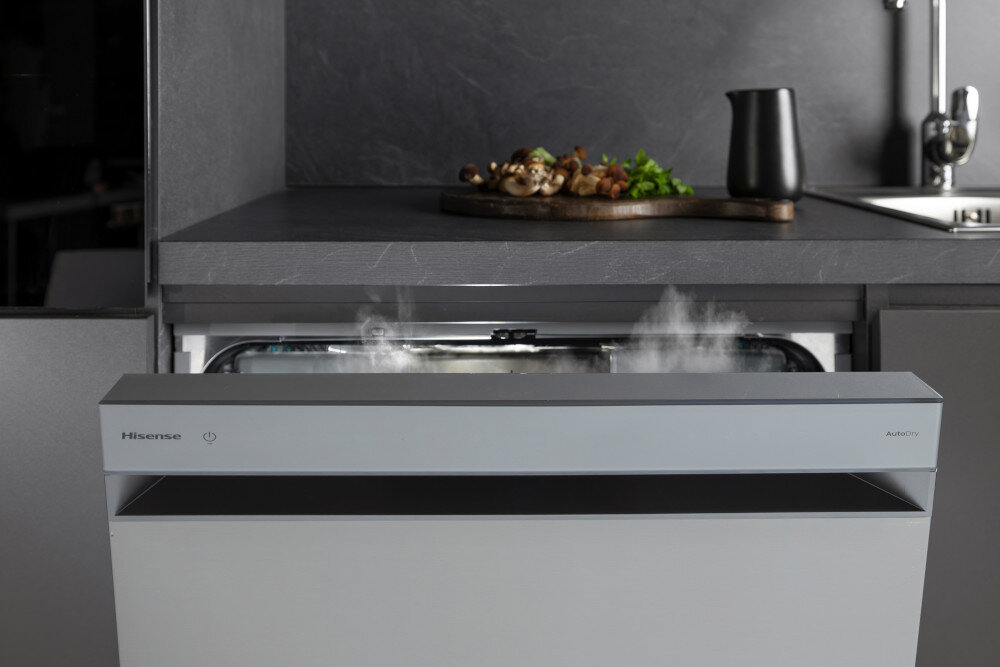 ZMYWARKA HISENSE HS693A90XAD Na zdjęciu otwarta zmywarka Hisense wysuwa się spod ciemnego blatu kuchennego, z którego unosi się para wodna z jej wnętrza. Nad urządzeniem stoi deska do krojenia z warzywami i pieczonymi szaszłykami oraz czarny dzbanek ustawiony przy zlewie automatyczne otwieranie drzwi naturalne suszenie