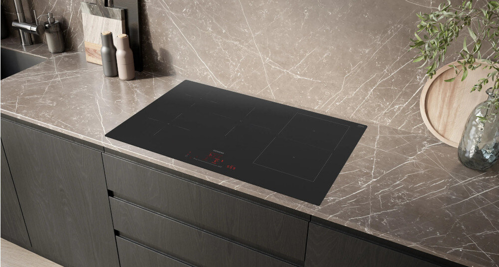 PŁYTA SIEMENS ED81AHWB1E 1.jpg przedstawia czarną, prostokątną płytę indukcyjną wbudowaną w jasny, marmurkowy blat kuchenny. Płyta ma cztery strefy gotowania oraz czerwony, dotykowy panel sterowania umieszczony z przodu. Otaczają ją ciemne, nowoczesne szafki oraz dekoracje kuchenne, co podkreśla elegancki charakter zabudowy