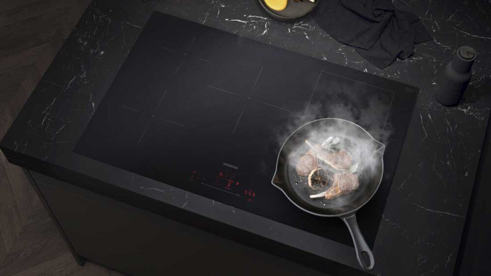 PŁYTA SIEMENS ED81AHWB1E 15.jpg pokazuje szeroką, czarną płytę indukcyjną Siemens zamontowaną w ciemnym, marmurkowym blacie kuchennym. Na jednym z pól grzewczych stoi patelnia z podsmażanymi kotletami jagnięcymi, z której unosi się para, a na dole płyty widać subtelnie podświetlony czerwony panel sterowania blokada rodzicielska płyta indukcyjna zabezpieczenie przed dziećmi bezpieczeństwo