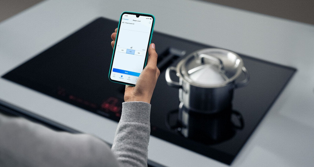 PŁYTA SIEMENS ED81AHWB1E 5.jpg pokazuje osobę stojącą przy czarnej płycie indukcyjnej, na której znajduje się garnek ze stalową pokrywką. W dłoni trzyma smartfon z otwartą aplikacją do zdalnego sterowania płytą, na ekranie widoczne są ustawienia poziomu mocy. Scena podkreśla wygodę kontroli gotowania za pomocą telefonu Home Connect Siemens zdalne sterowanie urządzeniami smart home WiFi inteligentne AGD sterowanie przez aplikację kuchnia Siemens smart living