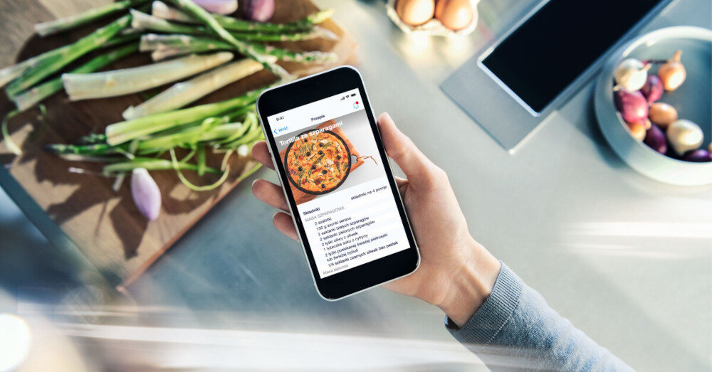 PŁYTA SIEMENS ED81AHWB1E 6.jpg przedstawia osobę trzymającą smartfon z wyświetlonym przepisem na tortillę ze szparagami. Na blacie obok leży deska z pociętymi szparagami i cebulką, a w tle widać inne składniki, co tworzy atmosferę gotowania z pomocą aplikacji kulinarnej przepisy Home Connect inteligentne gotowanie automatyczne ustawienia smażenia czujnik smażenia płyta indukcyjna Siemens smart kuchnia gotowanie bez wysiłku