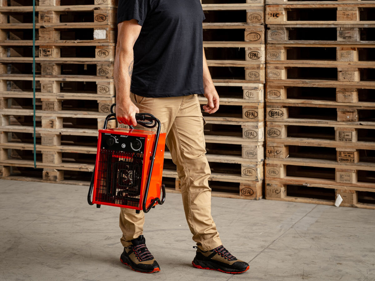 Nagrzewnica elektryczna DEDRA DED9922, uchwyt, lekkość, czystość, stabilność, Mężczyzna niesie czerwoną nagrzewnicę Dedra DED9922 w magazynie, palety w tle.