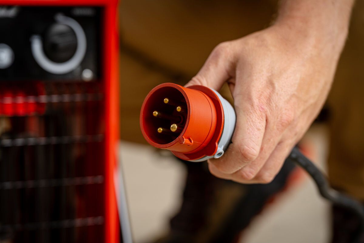 Nagrzewnica elektryczna DEDRA DED9922, 400V, trójfazowość, przemysł, stabilność, Czerwona wtyczka zasilająca 400V do nagrzewnicy DED9922, zbliżenie styków, ręka trzyma przewód.