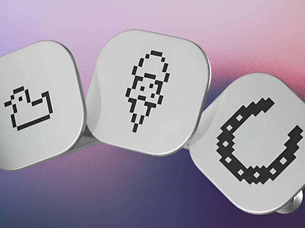 Odznaki LEDGER Nano Gen5 Pakiet Odznak 2 Zbliżenie na trzy małe, kwadratowe żetony lub przyciski Ledger Nano, ułożone w rzędzie, na rozmytym tle w odcieniach fioletu, różu i niebieskiego. Na żetonach widnieją czarne, pikselowe ikony: z lewej strony zarys kaczki, pośrodku lód w wafelku, a z prawej podkowa.