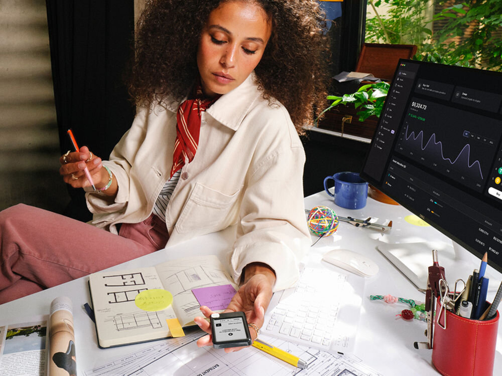 Portfel kryptowalut LEDGER Nano Gen5 Kobieta z kręconymi włosami w beżowej kurtce i różowych spodniach siedzi przy biurku pełnym dokumentów, notesów i akcesoriów biurowych. Trzyma w dłoni Ledger Nano z jasnym ekranem. Obok niej stoi monitor wyświetlający ciemny interfejs z wykresem kryptowalut.