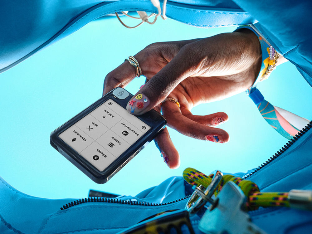 Portfel kryptowalut LEDGER Nano Gen5 Ujęcie z wnętrza turkusowej torebki lub plecaka, skierowane w górę, pod błękitne niebo. Ciemna dłoń, ozdobiona pierścionkami i kolorowym manicure, trzyma mały, czarny portfel sprzętowy Ledger Nano. Na ekranie widoczne są opcje Bitcoin, Ethereum, Solana, XRP, Security Key i Add app. W tle widać kolorową bransoletkę na nadgarstku i klucze na lince wewnątrz torby.