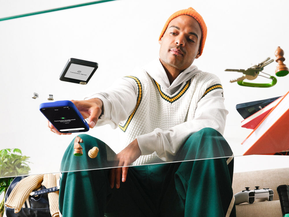 Portfel kryptowalut LEDGER Nano Gen5 Mężczyzna w pomarańczowej czapce i biało-żółtej kamizelce z dekoltem w serek wyciąga smartfon w kierunku widza. W tle, obok jego głowy, unosi się portfel sprzętowy Ledger Nano z ekranem pokazującym Transaction signed (Transakcja podpisana). Scena jest surrealistyczna, z unoszącymi się wokół przedmiotami, takimi jak szachy, klucze i kontroler do gier.