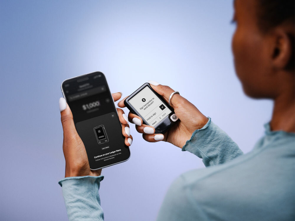 Portfel kryptowalut LEDGER Nano Gen5 Zbliżenie na dłonie trzymające dwa urządzenia: po lewej smartfon, po prawej portfel sprzętowy Ledger Nano. Kobieta (widoczna od tyłu) przygotowuje się do wysłania Bitcoina. Na ekranie telefonu widnieje kwota $1,000 i komunikat, aby kontynuować na urządzeniu Ledger Nano. Na ekranie portfela Ledger Nano widnieje prośba o podpisanie transakcji: Sign transaction to send bitcoin? z opcją Hold to sign.