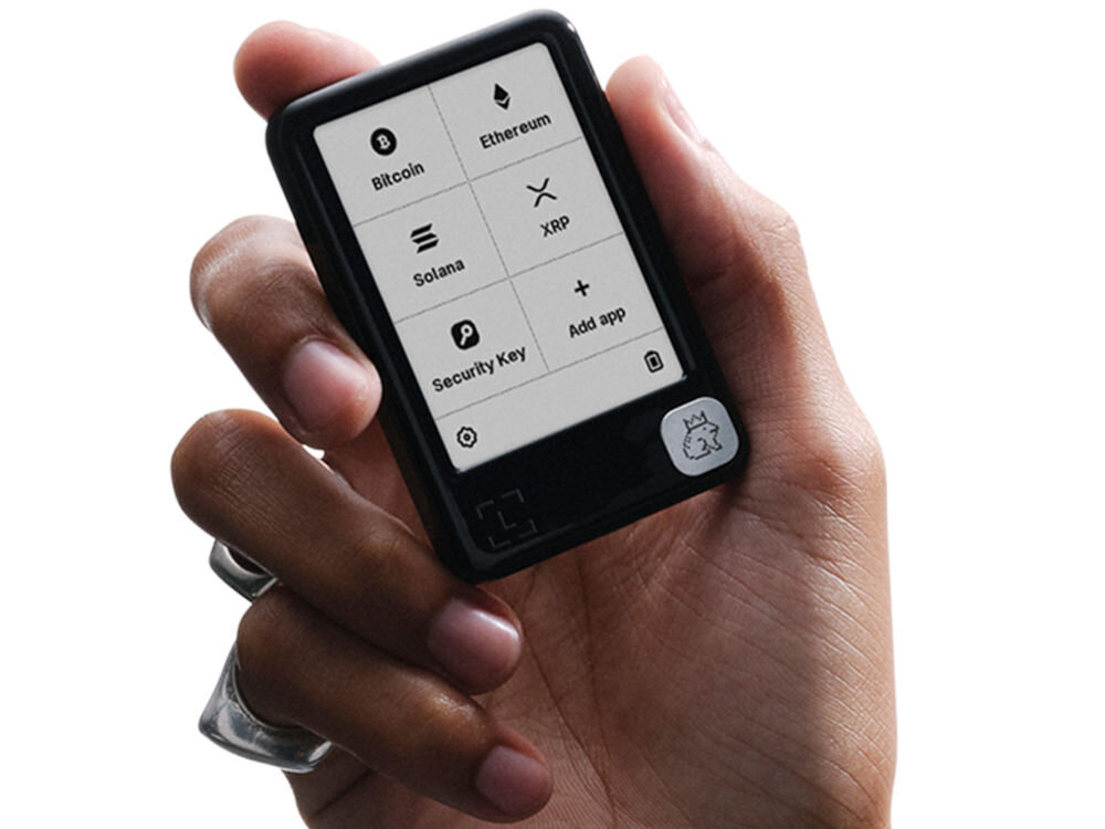 Portfel kryptowalut LEDGER Nano Gen5 Zbliżenie na dłoń trzymającą mały, czarny portfel sprzętowy Ledger Nano. Ekran urządzenia jest podświetlony i wyświetla menu główne z ikonami i nazwami popularnych kryptowalut: Bitcoin, Ethereum, Solana, XRP, a także opcje Security Key i Add app. U dołu ekranu widoczne są ikony ustawień i kosza na śmieci, a także mała ikonka smoka lub stworzenia w prawym dolnym rogu ramki urządzenia.