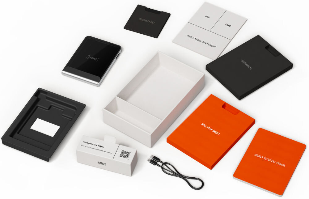 Portfel kryptowalut LEDGER Flex Widok rozłożonego zestawu i zawartości opakowania Ledger Stax. Wśród elementów znajdują się: sam portfel sprzętowy Ledger Stax, pudełka, kabel USB, instrukcje i dokumenty, w tym Recovery Sheet (arkusz odzyskiwania), SECRET RECOVERY PHRASE (tajna fraza odzyskiwania), oraz mała czarna karta z napisem RECOVERY KEY.
