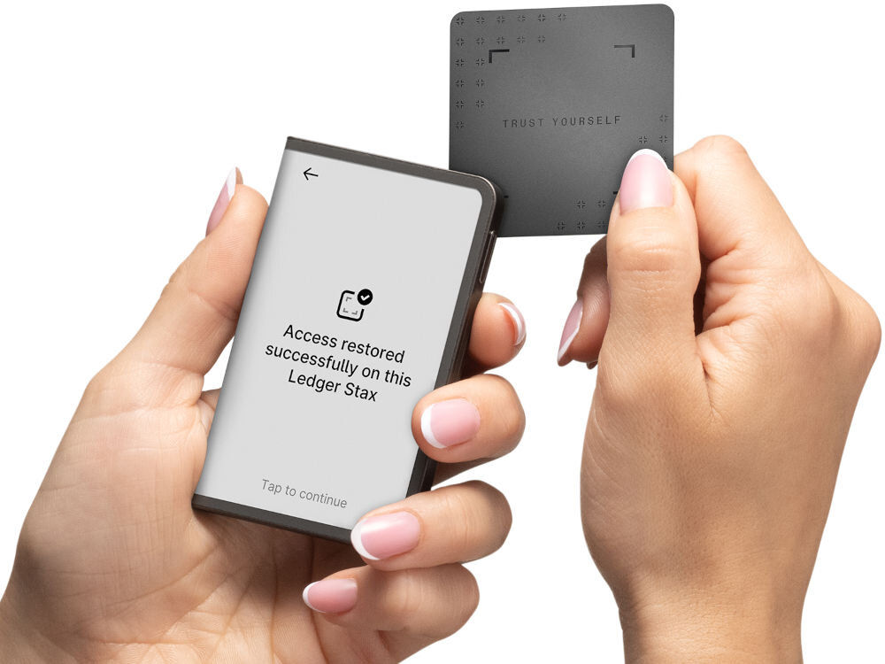 Portfel kryptowalut LEDGER Stax Crypto Hardware Wallet + Klucz odzyskiwania Dłonie trzymają portfel sprzętowy Ledger Stax i czarną kartę Ledger Recovery Card. Na ekranie Stax widnieje komunikat potwierdzający odzyskanie dostępu: Access restored successfully on this Ledger Stax oraz Tap to continue. Karta Recovery Card jest trzymana prawą ręką.