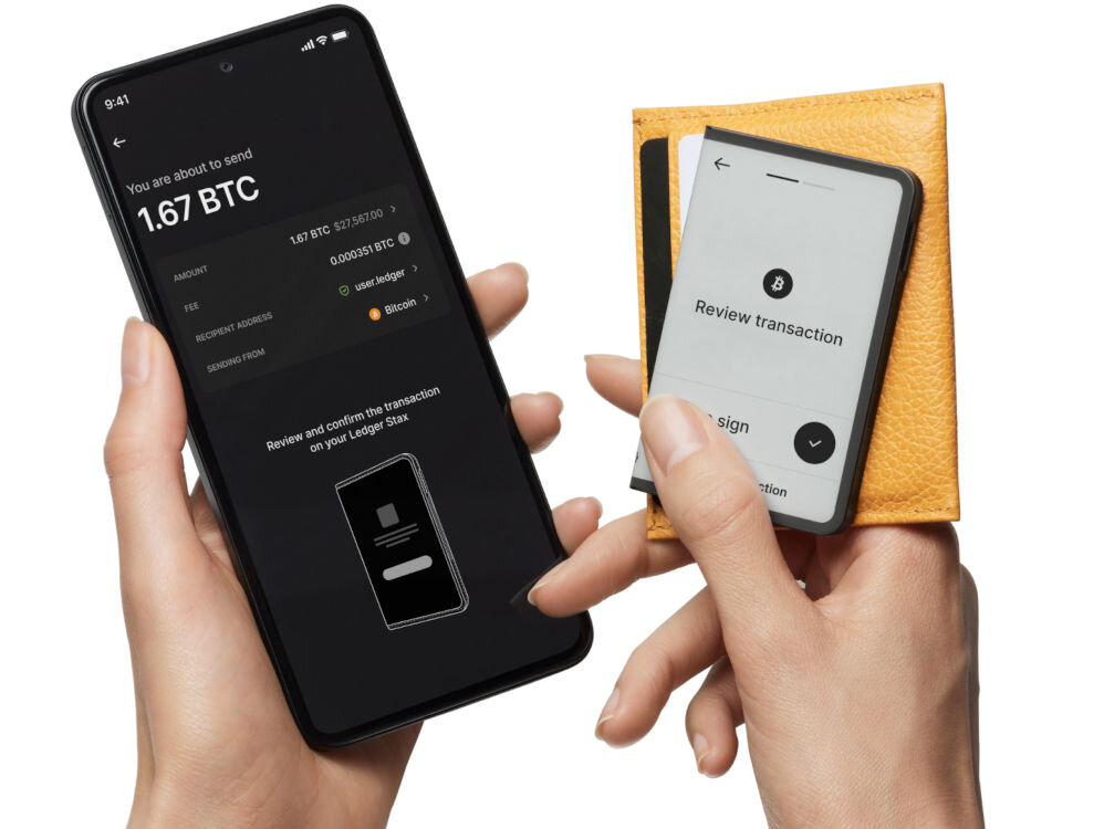Portfel kryptowalut LEDGER Stax Crypto Hardware Wallet + Klucz odzyskiwania Dłonie używają smartfona i portfela sprzętowego Ledger Stax do autoryzacji transakcji. Na ekranie smartfona widoczne jest podsumowanie transakcji wysłania 1.67 BTC z prośbą o przejrzenie i potwierdzenie na urządzeniu Ledger Stax. Ekran Ledger Stax, umieszczonego w żółtym skórzanym etui, wyświetla prośbę: Review transaction z ikoną Bitcoina.