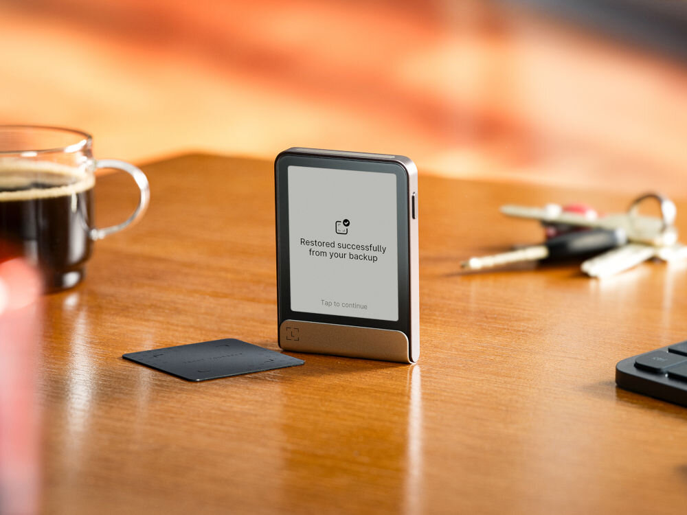 Portfel kryptowalut LEDGER Flex Ledger Stax stoi na drewnianym blacie obok szklanego kubka z kawą i pęku kluczy. Na ekranie urządzenia widnieje komunikat: Restored successfully from your backup (Pomyślnie przywrócono z kopii zapasowej) z opcją Tap to continue (Dotknij, aby kontynuować). Obok leży Ledger Recover Card.
