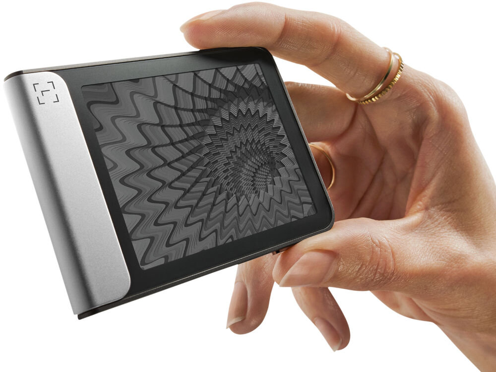 Portfel kryptowalut LEDGER Flex Zbliżenie na Ledger Stax trzymany w dłoni, pokazujące dynamiczną grafikę o hipnotycznym, falistym wzorze na ekranie e-ink (papier elektroniczny). Urządzenie jest zwrócone bokiem, eksponując jasny zielono-turkusowy element.