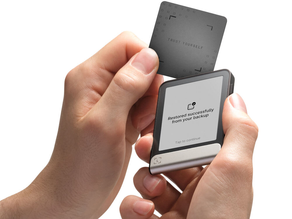 Portfel kryptowalut LEDGER Flex Ręce trzymają Ledger Stax oraz zbliżają do niego Ledger Recover Card (kartę do odzyskiwania). Na ekranie urządzenia widać komunikat: Restored successfully from your backup. Zielono-turkusowy pasek na dole ma animowany symbol potwierdzenia w kształcie nawiasów.