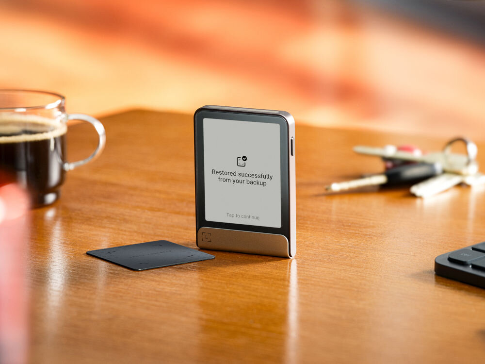 Portfel kryptowalut LEDGER Flex Ledger Stax stoi na drewnianym blacie obok szklanego kubka z kawą i pęku kluczy. Na ekranie urządzenia widnieje komunikat: Restored successfully from your backup (Pomyślnie przywrócono z kopii zapasowej) z opcją Tap to continue (Dotknij, aby kontynuować). Obok leży Ledger Recover Card.
