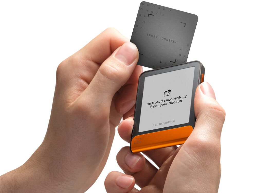 Portfel kryptowalut LEDGER Flex Ręce trzymają Ledger Stax oraz zbliżają do niego Ledger Recover Card (kartę do odzyskiwania). Na ekranie urządzenia widać komunikat: Restored successfully from your backup. Zielono-turkusowy pasek na dole ma animowany symbol potwierdzenia w kształcie nawiasów.