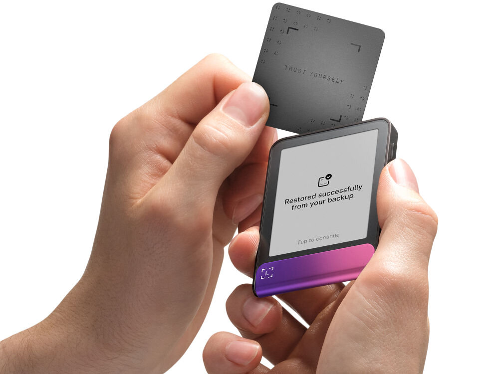 Portfel kryptowalut LEDGER Flex Ręce trzymają Ledger Stax oraz zbliżają do niego Ledger Recover Card (kartę do odzyskiwania). Na ekranie urządzenia widać komunikat: Restored successfully from your backup. Zielono-turkusowy pasek na dole ma animowany symbol potwierdzenia w kształcie nawiasów.