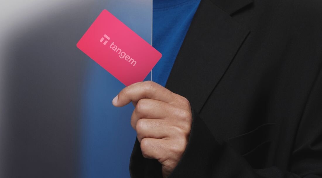 Osoba trzyma kartę Tangem w dłoni na tle przejścia z ciemnego do jasnego niebieskiego.  