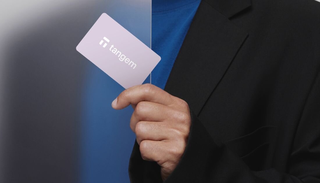 Osoba trzyma fioletową kartę Tangem w dłoni na tle przejścia z ciemnego do jasnego niebieskiego.  