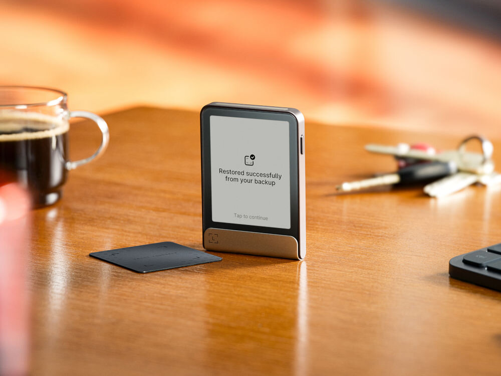 Portfel kryptowalut LEDGER Flex Ledger Stax stoi na drewnianym blacie obok szklanego kubka z kawą i pęku kluczy. Na ekranie urządzenia widnieje komunikat: Restored successfully from your backup (Pomyślnie przywrócono z kopii zapasowej) z opcją Tap to continue (Dotknij, aby kontynuować). Obok leży Ledger Recover Card.
