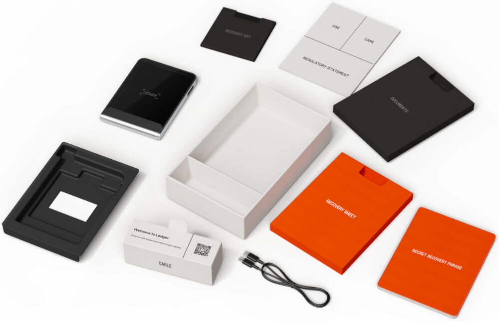 Portfel kryptowalut LEDGER Flex Widok rozłożonego zestawu i zawartości opakowania Ledger Stax. Wśród elementów znajdują się: sam portfel sprzętowy Ledger Stax, pudełka, kabel USB, instrukcje i dokumenty, w tym Recovery Sheet (arkusz odzyskiwania), SECRET RECOVERY PHRASE (tajna fraza odzyskiwania), oraz mała czarna karta z napisem RECOVERY KEY.