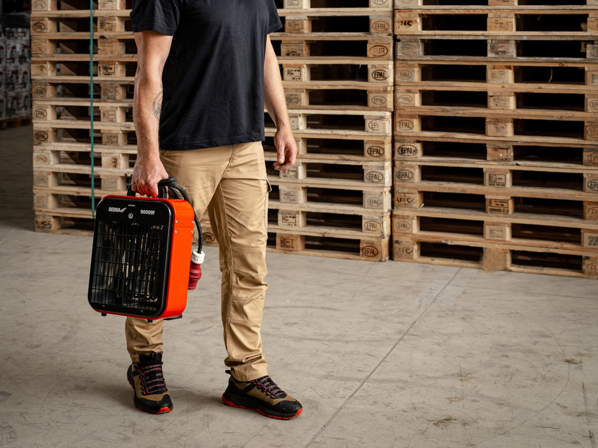 Nagrzewnica elektryczna DEDRA DED9924B, uchwyt, 6,5kg, ergonomia, prostota, Mężczyzna niesie nagrzewnicę Dedra DED9924B po magazynie, uchwyt, przewód zasilający.