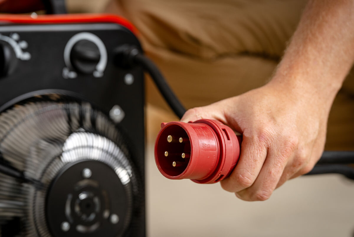 Nagrzewnica elektryczna DEDRA DED9924B, dogrzewanie, osuszanie, wentylacja, ciągłość, Dłoń trzyma dużą, czerwoną, przemysłową wtyczkę trójfazową od nagrzewnicy Dedra DED9924B.