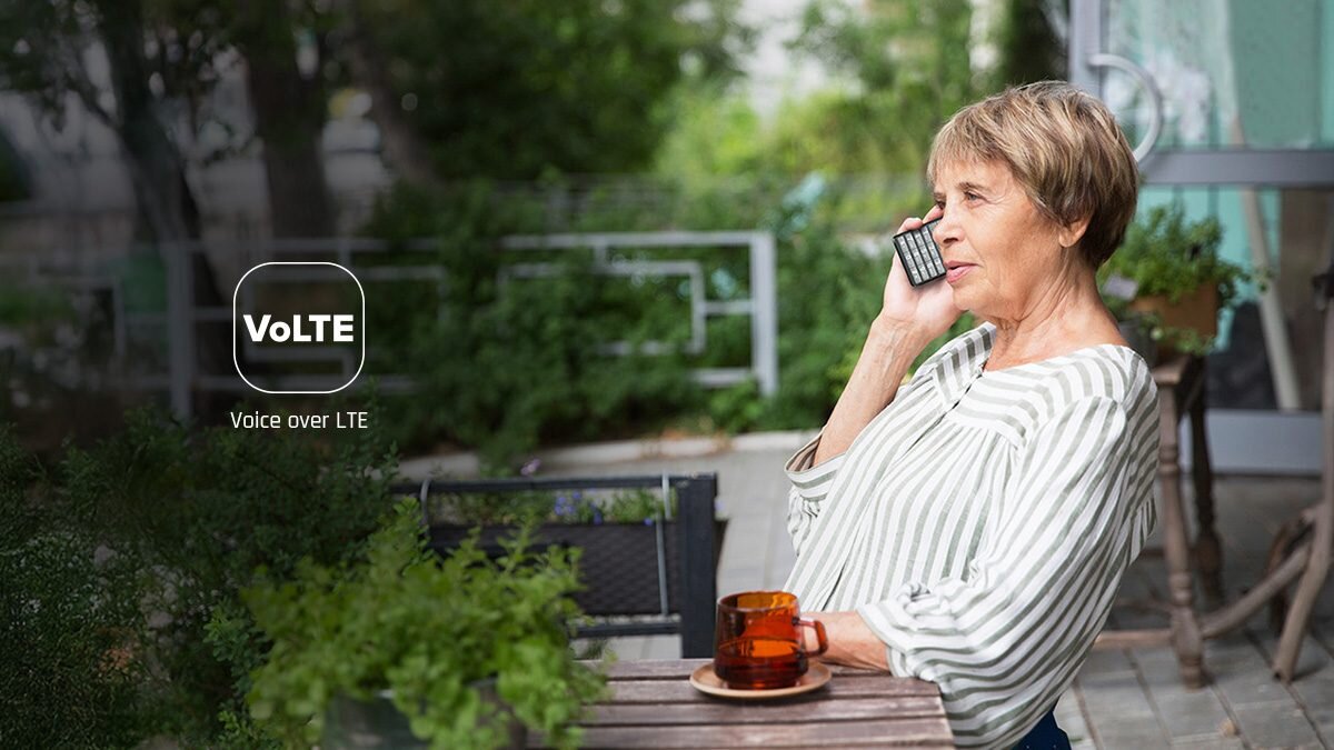 Starsza kobieta rozmawia przez telefon z klawiaturą na tarasie; po lewej ikonka „VoLTE”. Na stole szklanka z herbatą.