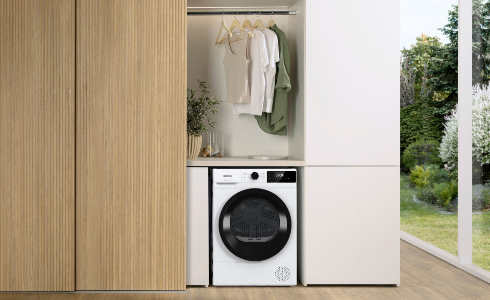 Na obrazie znajduje się suszarka w zabudowie, z widocznym logo \'gorenje\' na froncie. Nad suszarką wisi kilka ubrań na wieszakach, a obok stoi roślina w doniczce.