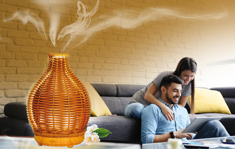 Na obrazie widoczny jest aromatyzer w kształcie wazonu z wikliny, z którego unosi się para. W tle znajduje się para siedząca na kanapie, przeglądająca tablet.