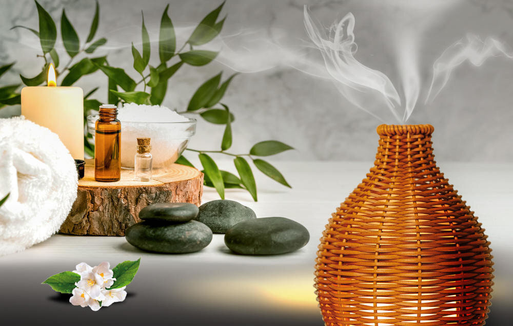 Na obrazie widoczny jest brązowy aromatyzer z wikliny, z którego unosi się para. W tle znajdują się kamienie, świeca, buteleczki z olejkami oraz liście roślin.